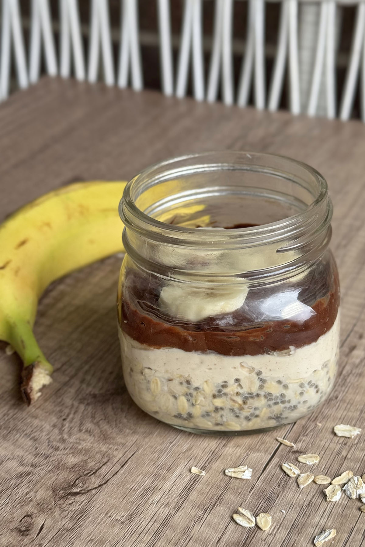 Protein-packed peanut butter chocolate oats breakfast jar, topped with banana slices with a banana on the background.